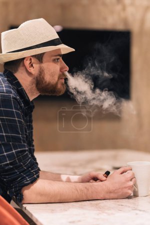 Photo for Young man in hat drinking coffee and smoking an electronic cigarette during break from work. Lifestyle concept. - Royalty Free Image