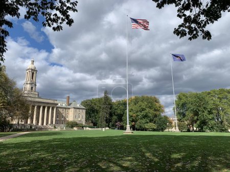Photo for State College, Pennsylvania, USA - October 7, 2023: Old main building at the campus of Pennsylvania State University in State College, PA with the Flag of the United States of America and the flag of Pennsylvania waving in front, framed by leaves. - Royalty Free Image