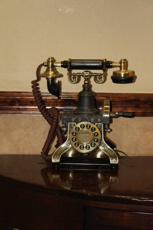 A nostalgic telephone with distinctive design features, such as rotary dials, ornate detailing in black and gold, telephone receiver and cable is standing on a table.