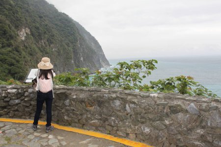 Foto de Hualien, Taiwán-18 de marzo de 2024: Mujer asiática con el pelo largo y oscuro abierto mira a la costa, con mochila y sombrero solar en la barandilla de roca en el acantilado de Quingshui. Océano Pacífico en Hualien, Taiwán. Costa tropical, isla. - Imagen libre de derechos