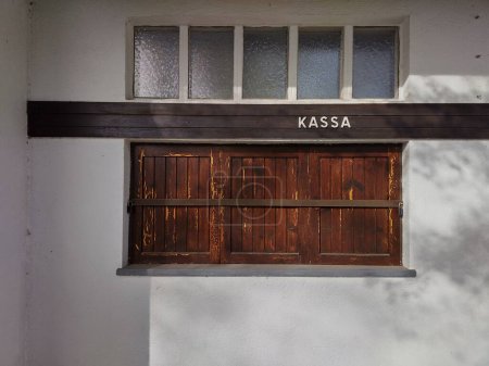 Window on white wall barricaded with wooden board. Closed checkout,  shut entrance. German word Kassa is written above, which means register. Abandoned entrance to a public pool in winter time. Closed for the season