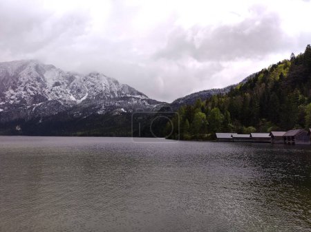 Panoramic view at lake Altaussee in Austria, surrounded by snowy mountain peaks. Lined up boathouses at waterfront and green trees. Cloudy day in spring.