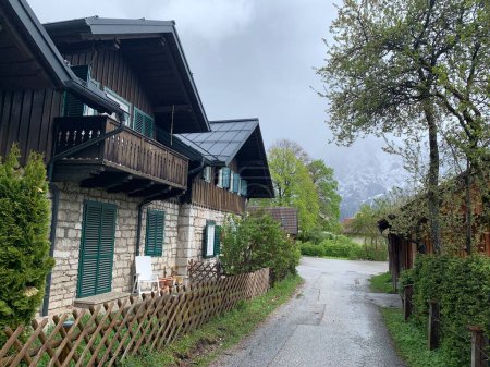 Foto de Altaussee, Austria, April 20, 2024: Traditional small houses with wooden details, green shutters, porch and fence on small road in Austria. Small village in the Alps. Cloudy day in spring, snowy - Imagen libre de derechos