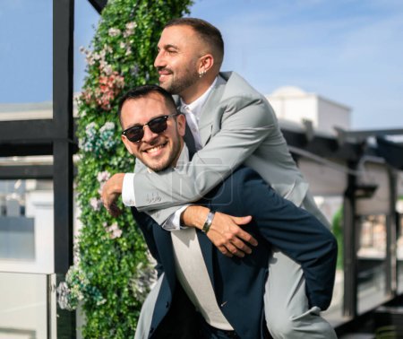 Couple homosexuel nouvellement marié. L'un d'eux tient l'autre sur le dos, très heureux du mariage. À l'extérieur, avec la lumière du soleil. Espace de copie.