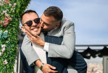 Couple d'hommes nouvellement mariés. Concept gay. L'un d'eux tient l'autre sur son dos pendant qu'il reçoit un baiser sur la joue. À l'extérieur, sous un gui.