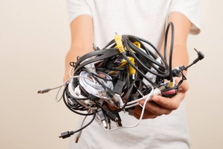 Hands holding pile of tangled old smart technology wires, used charging cables and cords. E-waste, planned obsolescence, electronic donation, disposal of electronic waste, recycling concept.