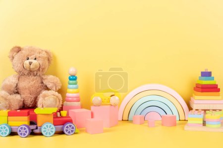 Kolekcja edukacyjnych zabawek dla dzieci. Miś, drewniana tęcza, pociąg, układanie różowych kostek piramidy na żółtym tle. Widok z przodu.