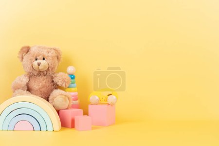 Educational kids toys collection. Teddy bear, wooden rainbow pink cubes and colorful balls on yellow background. Front view.