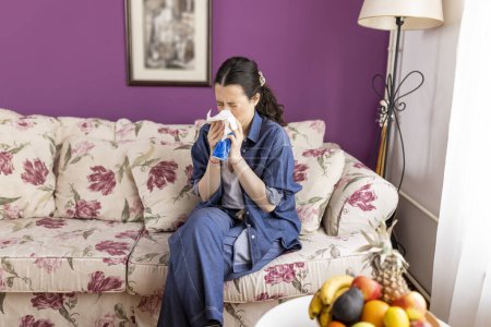 Sick young woman sitting on the sofa holding tissue handkerchief blowing running nose feels unwell unhealthy, girl having symptoms of chronic sinusitis disease, seasonal allergy or cold fever flu 