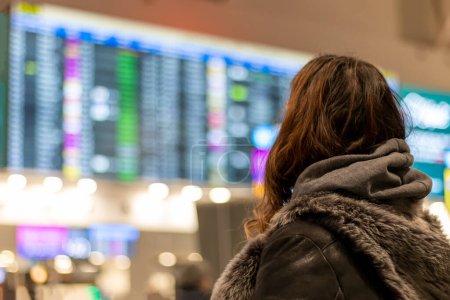 Téléchargez les photos : Vue de l'arrière d'une touriste féminine debout à l'aéroport et regardant le tableau de bord qui indique l'horaire des vols - en image libre de droit