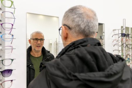 Un homme mûr en train d'essayer des lunettes dans la vitrine des lunettes et en regardant le miroir. Homme heureux de choisir des lunettes au magasin d'optique. Soins de santé, vision et concept de vision.
