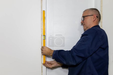 A worker's hands with  spirit level against frame of  the door. An experienced mature carpenter  installing interior door with a wooden frame at new  apartment. 