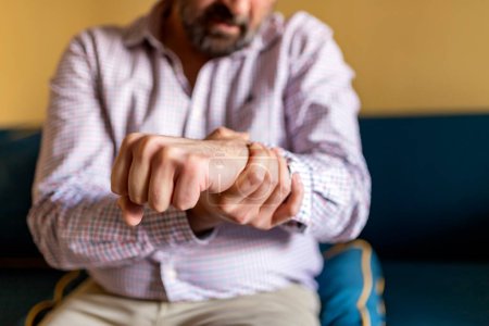 Mature Bearded Man Massaging His Painful Wrist. Man Suffering From Wrist Pain At Home, Sitting on the Sofa
