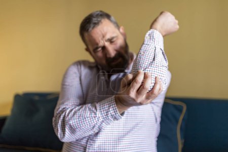Téléchargez les photos : Homme barbu mature massant son coude douloureux. Homme souffrant de douleur au coude à la maison, assis sur le canapé - en image libre de droit