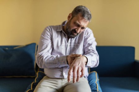 Reifer bärtiger Mann massiert sein schmerzhaftes Handgelenk. Mann leidet zu Hause unter Handgelenkschmerzen, sitzt auf dem Sofa