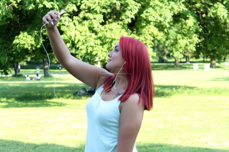 Photo for Seductive young redhead woman taking a selfie photo in public park while listening to music - Royalty Free Image