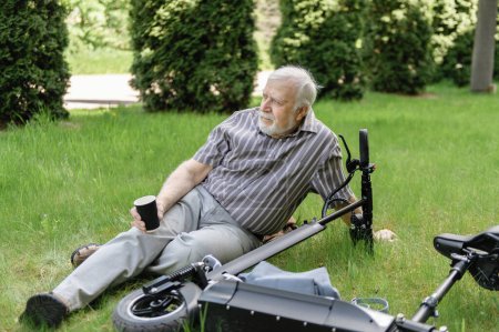 Foto de Un hombre positivo y anciano fue a dar un paseo por el parque, se detuvo y se acostó a descansar en la hierba junto a un scooter eléctrico. Verano, día soleado. El estilo de vida saludable de los ancianos. - Imagen libre de derechos