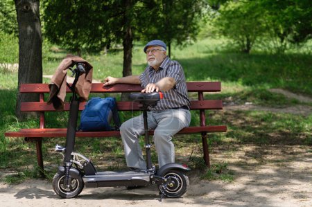 Photo for A positive, elderly man went for a walk to the park, stopped and sat down to rest on a bench next to an electric scooter. Summer, sunny day. Healthy lifestyle of the elderly. - Royalty Free Image
