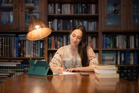 Téléchargez les photos : Femme écrivant sur papier avec bibliothèque en arrière-plan - en image libre de droit