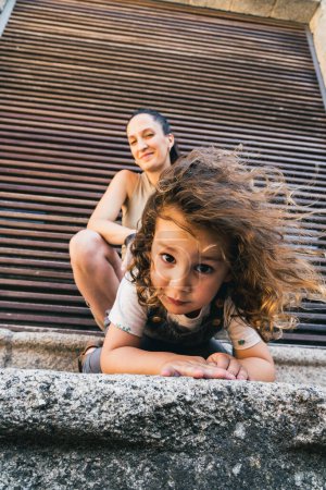 Téléchargez les photos : Petit garçon avec sa mère, accroupi en regardant la caméra tandis que les cheveux souffle dans le vent, voyageant à travers la ville - en image libre de droit