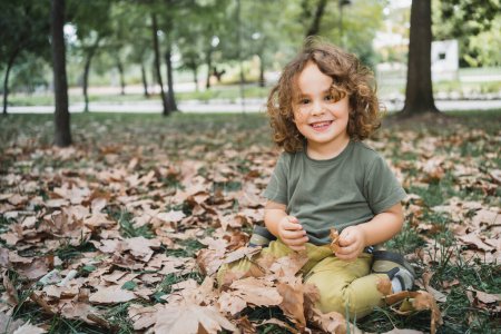 Foto de Retrato de niño feliz jugando en la hierba con hojas secas en un parque en un día soleado mirando a la cámara - Imagen libre de derechos
