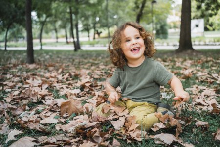 Foto de Retrato de niño feliz jugando en la hierba con hojas secas en un parque en un día soleado mirando hacia los lados - Imagen libre de derechos