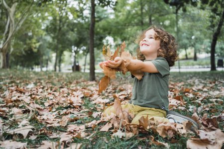 Foto de Retrato de niño feliz jugando en la hierba con hojas secas en un parque en un día soleado - Imagen libre de derechos
