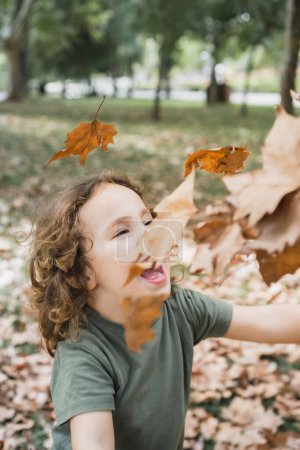 Photo for Portrait of happy boy playing on the grass with dry leaves in a park on sunny day, vertical - Royalty Free Image