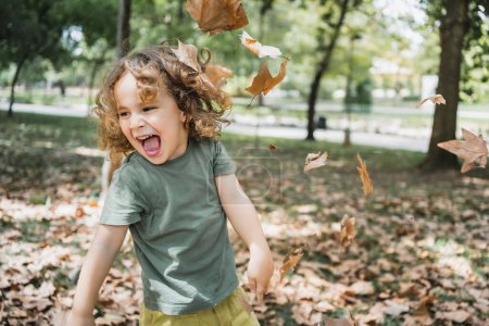 Foto de Retrato de niño feliz jugando en la hierba con hojas secas en un parque en un día soleado - Imagen libre de derechos