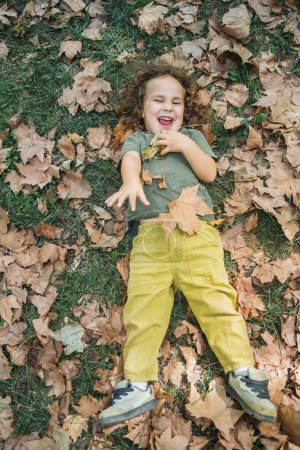 Téléchargez les photos : Enfant heureux couché jouant sur l'herbe avec des feuilles sèches, pleine photo du corps - en image libre de droit