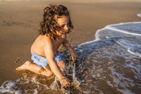 Téléchargez les photos : Petit garçon jouant avec l'eau sur le bord de mer assis sur le sable de la plage - en image libre de droit