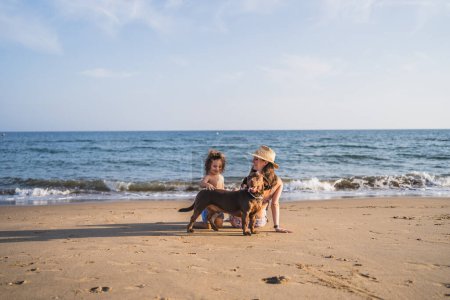 Téléchargez les photos : Mère et fils face à la mer assis jouer avec un chien - en image libre de droit