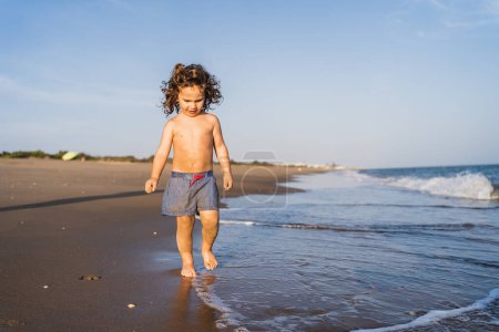 Foto de Niño pequeño caminando descalzo a lo largo de la playa en traje de baño - Imagen libre de derechos