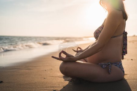 Téléchargez les photos : Femme enceinte méditant assis sur le rivage de la plage regardant vers la mer - en image libre de droit