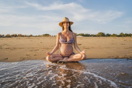 Téléchargez les photos : Femme enceinte méditant assis sur le rivage de la plage regardant vers la mer - en image libre de droit