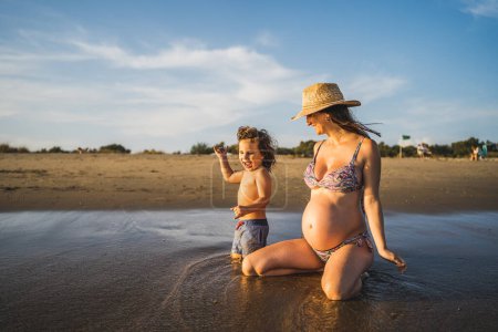 Téléchargez les photos : Mère enceinte jouant avec son fils sur le bord de la mer avec des vagues et du sable, lumière du soir - en image libre de droit