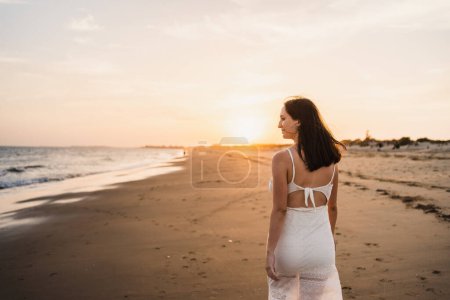 Téléchargez les photos : Belle femme heureuse marchant le long du rivage de la plage regardant vers la mer vêtue de blanc - en image libre de droit