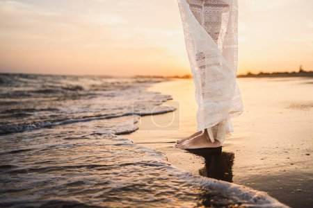 Foto de Piernas de una mujer vestida de blanco en la playa empapando sus pies en el agua del mar - Imagen libre de derechos