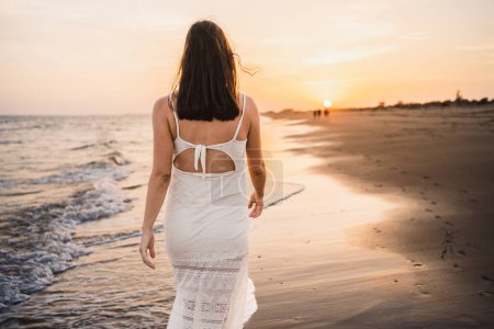 Photo for Beautiful woman walking along the beach shore in a nice soft dress in the sunset light - Royalty Free Image