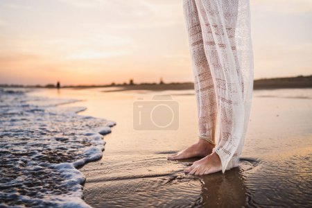Téléchargez les photos : Pieds de belle femme en belle robe blanche sur le rivage de la plage mouillée par les vagues - en image libre de droit