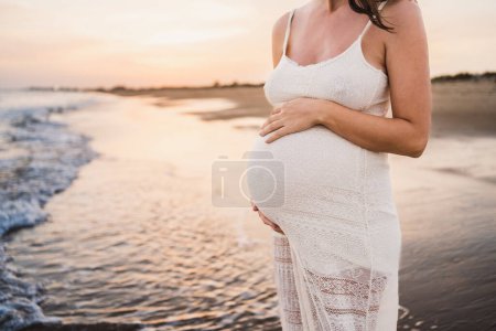 Téléchargez les photos : Portrait de belle femme enceinte en robe blanche, au bord de la mer avec lumière du coucher du soleil - en image libre de droit