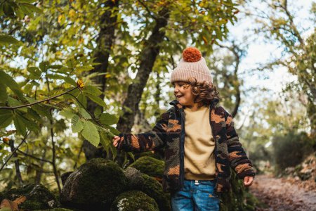 Photo for Portrait of fashionable little boy with hat playing happily on a forest path while teaching his parents about plants - Royalty Free Image