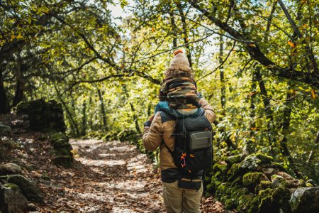 Téléchargez les photos : Photographe père avec sac à dos et son fils à la mode avec chapeau sur les épaules, heureux, marchant dans une forêt - en image libre de droit