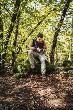 Photo for Young and beautiful photographer resting on a stone in the forest after walking and photographing the wonders of the environment, while looking at the photographs in the camera, vertical - Royalty Free Image