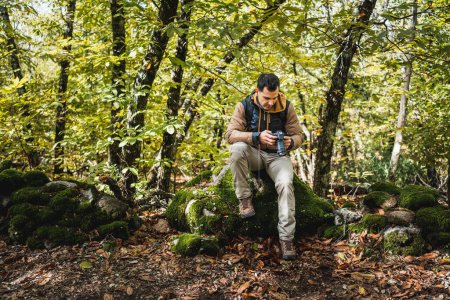 Foto de Joven y hermoso fotógrafo descansando sobre una piedra en el bosque después de caminar y fotografiar las maravillas del medio ambiente, mientras mira las fotografías en la cámara - Imagen libre de derechos