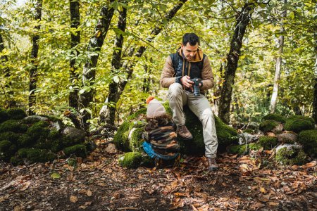 Foto de Fotógrafo padre con mochila y su hijo de moda con sombrero descansando sentado en una piedra en el bosque después de caminar por el camino - Imagen libre de derechos
