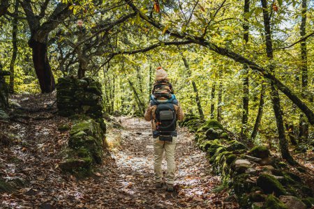 Foto de Fotógrafo padre con mochila y su hijo de moda con sombrero en los hombros, feliz, caminando por un bosque - Imagen libre de derechos