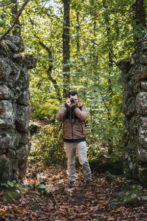 Foto de Foto del joven y hermoso fotógrafo tomando una foto de la vieja puerta de piedra en el bosque, mirando a la cámara, foto vertical - Imagen libre de derechos