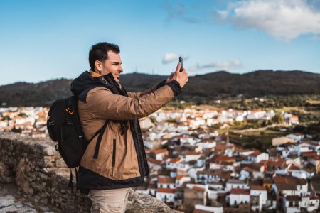 Photo for Young happy photographer man traveling sightseeing in a small town, taking a photo with his cell phone of the panoramic view of the town and the mountains, from a castle on a sunny day - Royalty Free Image
