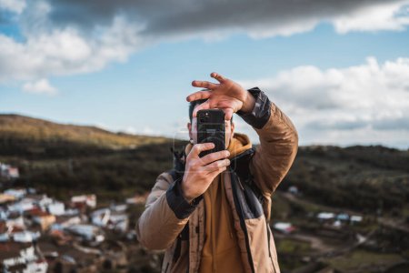 Photo for Young happy photographer man traveling sightseeing in a small town, taking a photo with his cell phone of the panoramic view of the town and the mountains, from a castle on a sunny day - Royalty Free Image